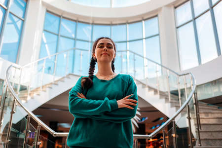 Smiling woman with pigtails stands on the steps, crossed arms and looks at the camera. In the background, there are windows and sunlight. Hero view.の写真素材