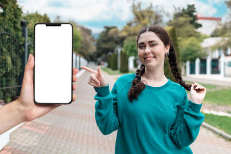 A smiling beautiful woman stands on the street and points to the side. Hand holding a smartphone with a white screen, mock up. Copy space. The concept of online communication.の写真素材