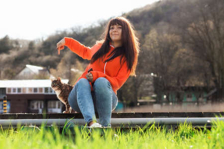 A smiling female volunteer sits on a bench near a stray hungry cat. A shelter worker takes care of the street animals. Concept of day of protection and care of stray animals.の写真素材