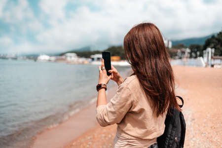 Girl with backpack takes a photo of the sea. Rear view. In the background there is a sandy beach.