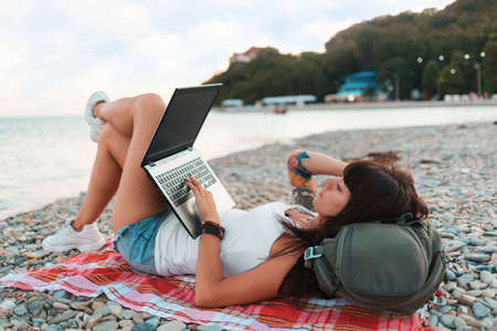 Summer vacation. A woman with a tattoo on her arm lies on the beach on the sand and works on a laptop. Side view. The concept of remote work and freelancing.の写真素材