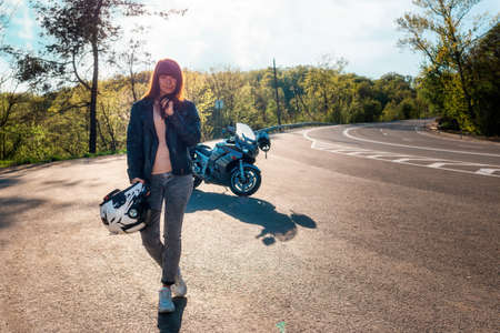 A young smiling woman in a leather jacket poses against the background of a motorcycle by the road. Motorcycle travel concept.の写真素材