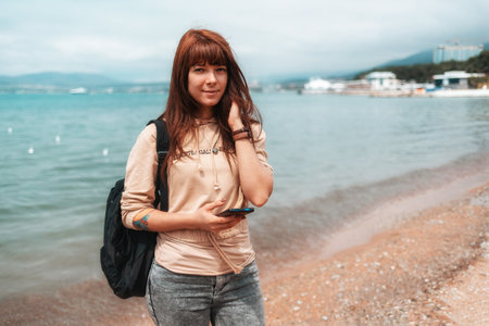 Vacation. Portrait of a cute smiling girl with a backpack and smartphone in her hand, posing on the beach.の写真素材
