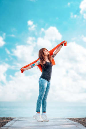 A young happy woman reaches up with her hand. Vertical orientation. Copy space. In the background, the blue sky with clouds and the ocean. Concept of freedom and carefree.の写真素材