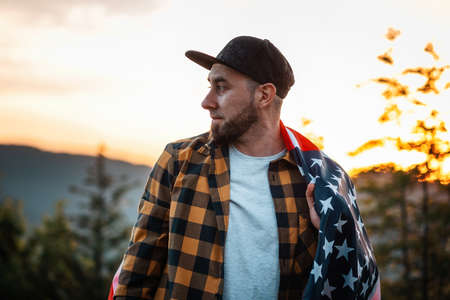 Portrait of patriotic young bearded man stand with an American flag at shoulders. Sunset in the background. The concept of American Independence day.の写真素材