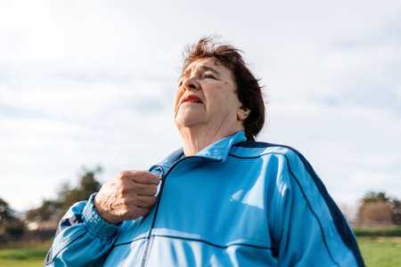 Portrait of an old woman buttoning up a sports jacket. In the background, the sky and the park. Sports Lifestyle concept.の写真素材