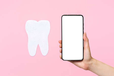 A female's hand holds a smartphone with a white screen, next to a tooth cut out of felt. Pink background. Mock up. The concept of modern stomatology.の写真素材