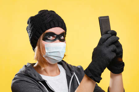 New normal. A female hacker wearing a black hat, gloves and a protective mask holds a mobile phone...