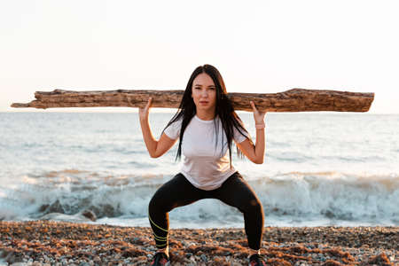 The concept of sport. Young brunette squats with a log on her shoulders. In the background, the sea...