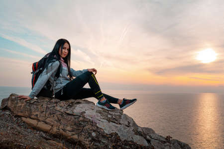 A young woman with a backpack on her back, sitting on the edge of a cliff. In the background, the sea and the sky at sunset. Copy space. Concept of Hiking and outdoor activities.の写真素材