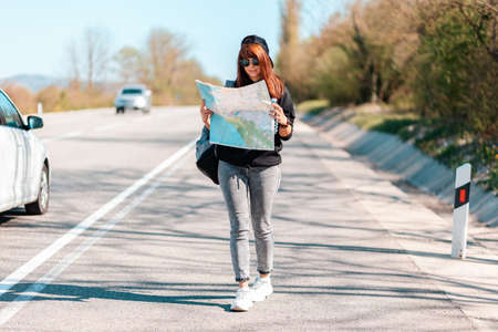 Hitchhiking trips. A young hipster woman in a cap and sunglasses, holding a paper map. In the background, a road with driving cars is blurred. The concept of local travel.の写真素材