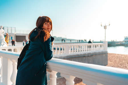 Vacation at the sea. A smiling woman leaning on a balustrade and looking at the camera. Rear view....