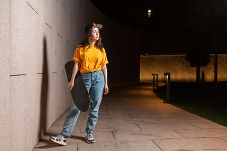 Caucasian young woman stand with a skateboard at her hands, posing on a night street. Copy space. The concept of youth subculture.の写真素材