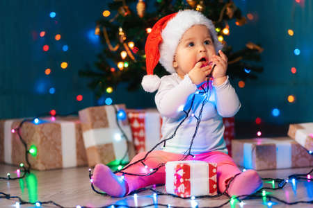 Portrait of a baby child in a Christmas hat, chewing festive lights. Indoor. The concept of New Year holidaysの写真素材