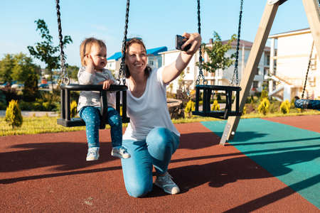 A young smiling mother takes a selfie with her baby sitting on a swing. Playground in the background. Summertime vacations.の写真素材