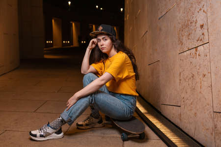 A serious teenage girl sits on a skateboard, posing on a night street. Outdoor. The concept of youth...