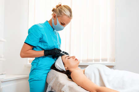 Woman getting injection at her forehead in the cosmetology salon. Cosmetologist in latex gloves and protective mask makes with syringe injects of a hyaluronic acid. Concept of plastic surgery and face liftingの写真素材