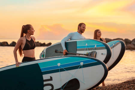 Summertime. Group of people walking on the beach with sup boards. In the background, the ocean and sunset. Copy space. Surfing.の写真素材
