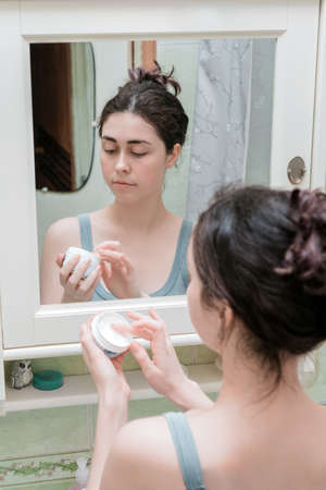 a young girl standing in front of a mirror holding a jar of cream in her hands for further cosmetic use.の写真素材