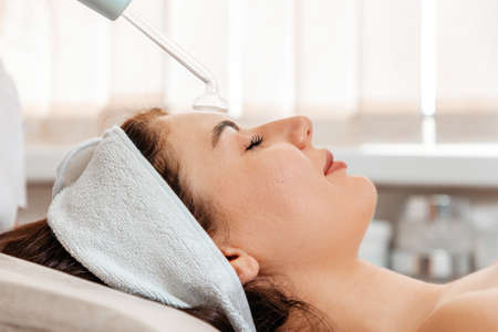 Darsonval cosmetology apparatus. Beautician cleans the client's forehead using a electric device. Close up. Professional rejuvenation procedure in salon.の写真素材