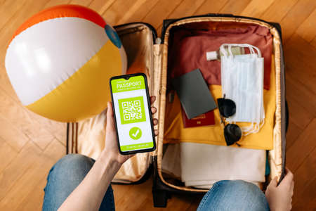 A woman holds a smartphone with an immune digital health passport in her hand. A negative test for the virus is covid-19. Flat lay. Close-up of a hand and a suitcase with clothes and virus protection equipment.の写真素材