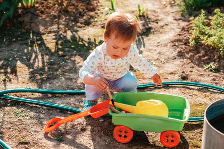 A cute toddler is playing with a plastic toy cart, putting gardening tools in it. Young assistant in...
