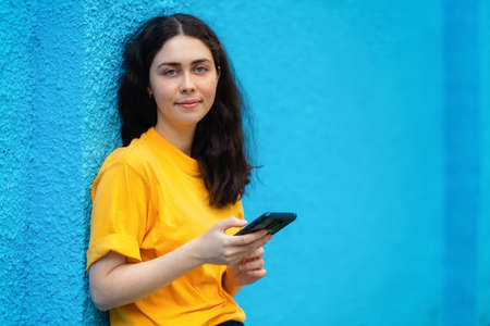 Portrait of a young smiling woman using a smartphone. Online surfing concept. Blue wall on the background. Copy space.の写真素材
