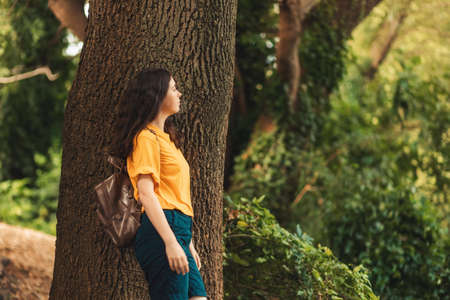 A lonely young Caucasian woman stands leaning on the trunk of a large tree and looks to the right side. In the background is a green summer forest. The concept of travel and psychology.の写真素材