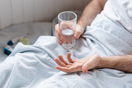 A man is lying in bed with a glass of water and holding a pill in the palm of his hand. Top view. Treatment concept.の写真素材