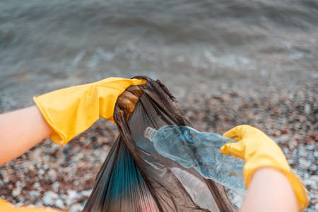 Close up of volunteer's hands in rubber gloves puts a dirty plastic bottle in bag with garbage. Ocean's water on the background. First-person view. The concept of envirinmental pollution.の写真素材
