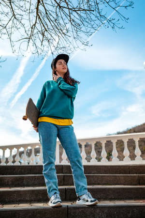 A young Caucasian woman with a skateboard in her hand stands on the steps. In the background, blue sky and trees. Bottom view. Copy space. Concept of sports lifestyle and street culture.の写真素材