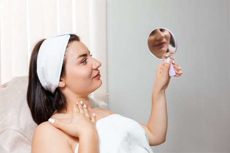 Plastic surgery. A young woman looks in the mirror during a procedure in a cosmetology salon. Concept of professional skin care.の写真素材