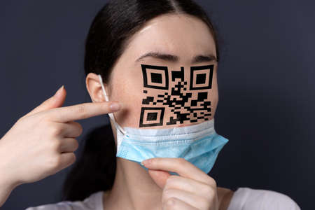 New reality. Close-up portrait of a woman taking off her mask and pointing at a face with a qr code....