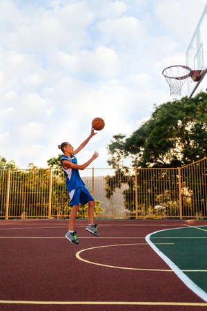 Basketball. A Teenage boy in blue sportswear jumps and throws a ball into a basketball Hoop. Sports field in the background. Cloudy sky. Side view. Copy space. Concept of sports games.の写真素材