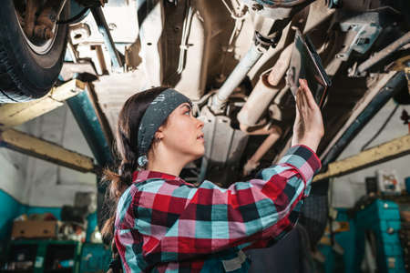Portrait of a young female mechanic in uniform who uses a tablet to diagnose a car, standing under a car on a lift. Bottom view.の写真素材