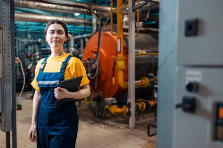 Portrait of a young smiling female worker in a work uniform and with a tablet in her hand. In the background is a boiler room. The concept of industrial production.の写真素材
