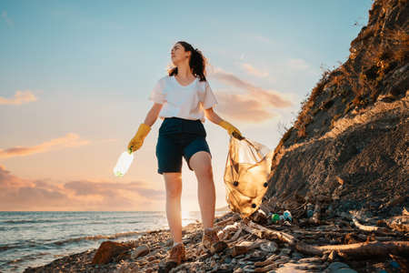 A female activist stands with a plastic bag and a bottle in her hand against the background of the sunset. Bottom view. Cleaning of the coastal zone and wild beaches. The concept of Earth Day.の写真素材