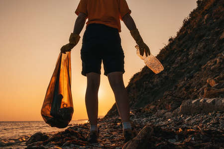 Zero waste. Silhouette of a volunteer holding a plastic bottle and posing on the beach. Sunset in the background. The concept of Earth Day and environmental conservation.の写真素材