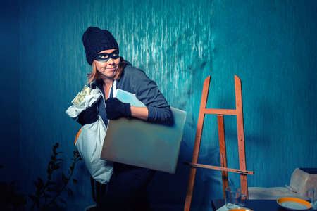 A masked female robber is carrying a stolen bag of money and a painting. The background is dark blue. The concept of theft and crime.の写真素材