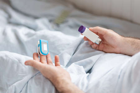 World Asthma Day. Patient lying at bed with pulse oximeter and holding a inhaler. Close up of hands....
