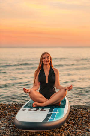 Yoga sup board. A young beautiful woman meditates on a surf board. In the background, the ocean, the horizon and the sunset. Recreation at the beach.の写真素材