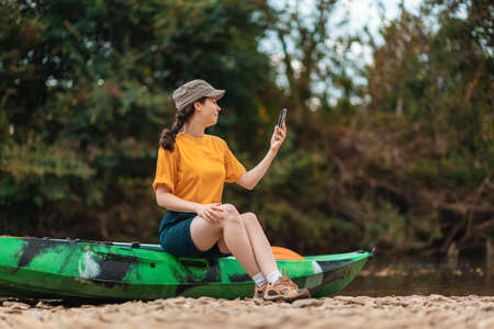 Kayaking. A young pretty woman in cap is sitting on a kayak and using a phone. The concept of GPS and internet navigation.の写真素材