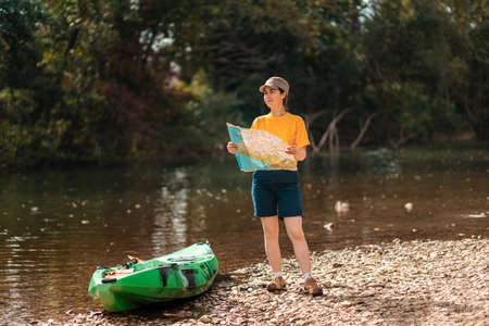 Young smiling woman holding a paper map and cheking the route. Kayak and river on the background. The concept of the World Tourism Day.の写真素材