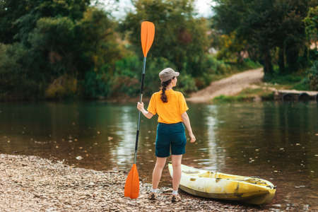 World Tourism Day. A young happy woman stands with an oar in her hand near the kayak. Back view. The concept of kayaking and outdoor activitiesの写真素材