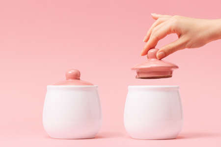 Two white porcelain pots with a pink lid on a pink background. Female hand removes the lid from the right side jar. Copy space. Concept of Breast Cancer Awareness Month.の写真素材