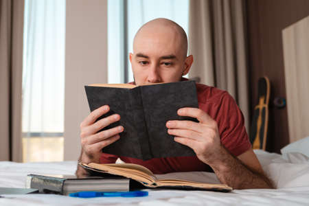 A bald caucasian man lies on the bed and reads a book. Home interior. The concept of learning and reading.の写真素材