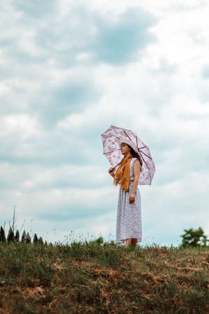 A young woman in a straw hat, dress, with an umbrella in her hands is standing on a hill. Cloudy sky...
