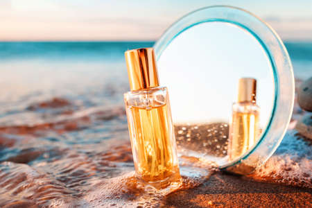 A golden glass perfume bottle with mirror on a sandy beach in surf sea. Close-up. Copy space. Perfume advertising concept.の写真素材