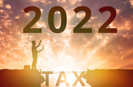 New Year 2022. The silhouette of a businessman with a laptop in his hand, pointing at the numbers in the sky. A bridge in the form of the word Tax. The sunset sky is on the background. The concept of changing the calendar year and calculating the annual taxの写真素材