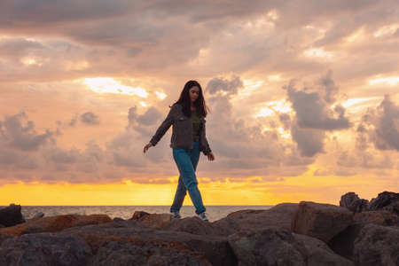 A young woman walks alone on the sea rocks. The dramatic sunset in the background. The concept of psychology and mental health.の写真素材